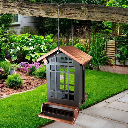 A hanging bird feeder with a dark gray body and copper-colored roof and tray, suspended from a tree branch in a landscaped garden with grass, plants, and a patio visible

perch & weather