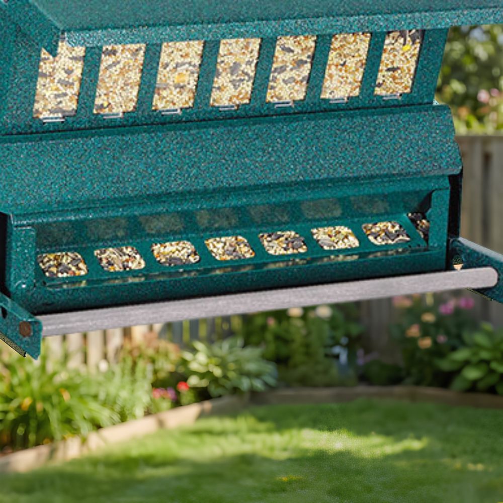 A close-up of a green bird feeder with clear seed windows and a metal tray below, shown outdoors in a garden with grass and plants blurred in the background

perch & weather
