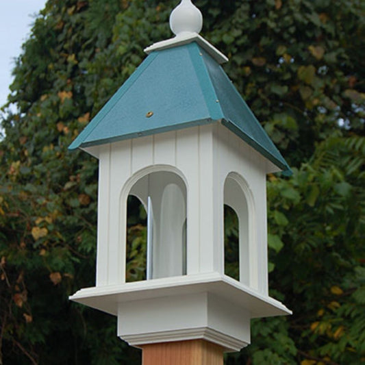 A white wooden bird feeder pole cap with a teal green pointed roof and white finials, mounted on a wooden post against a background of green trees

perch & weather